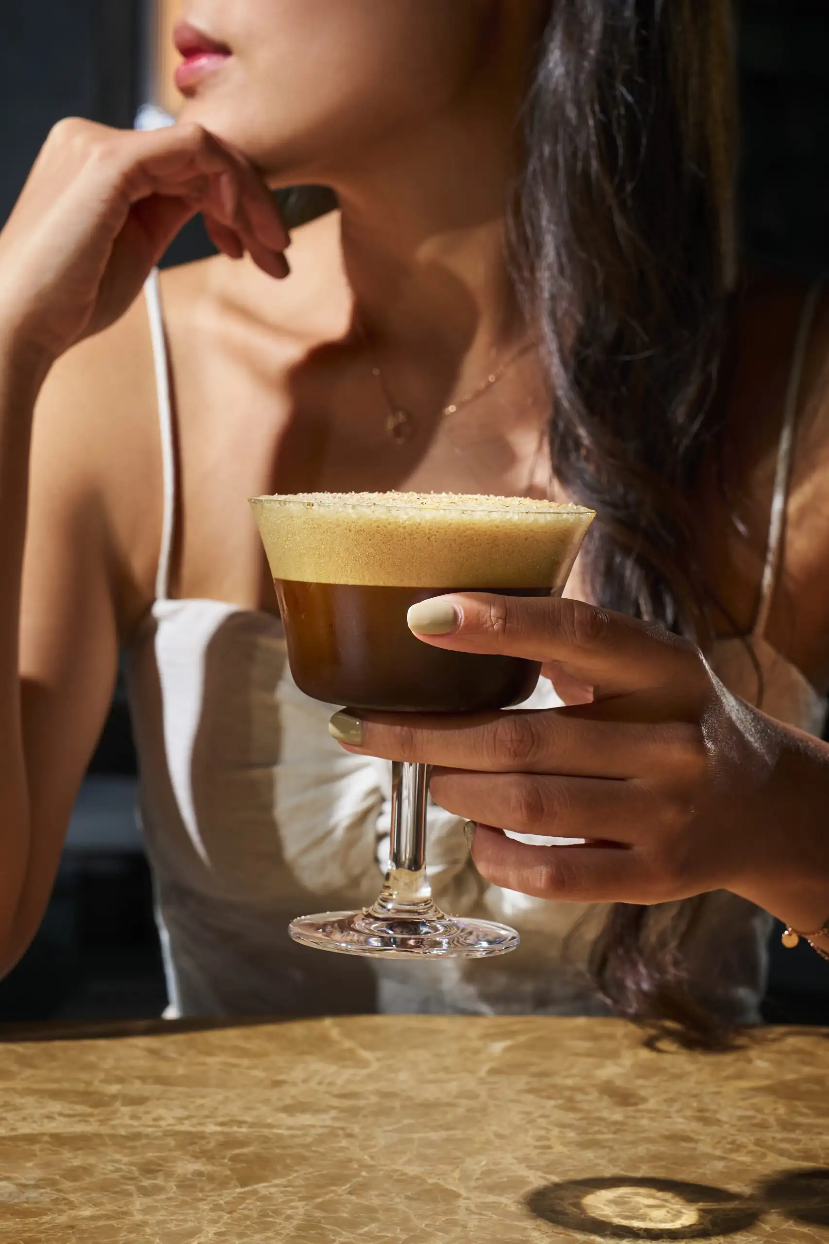 Woman holding a frothy espresso martini cocktail in a dimly lit bar setting.