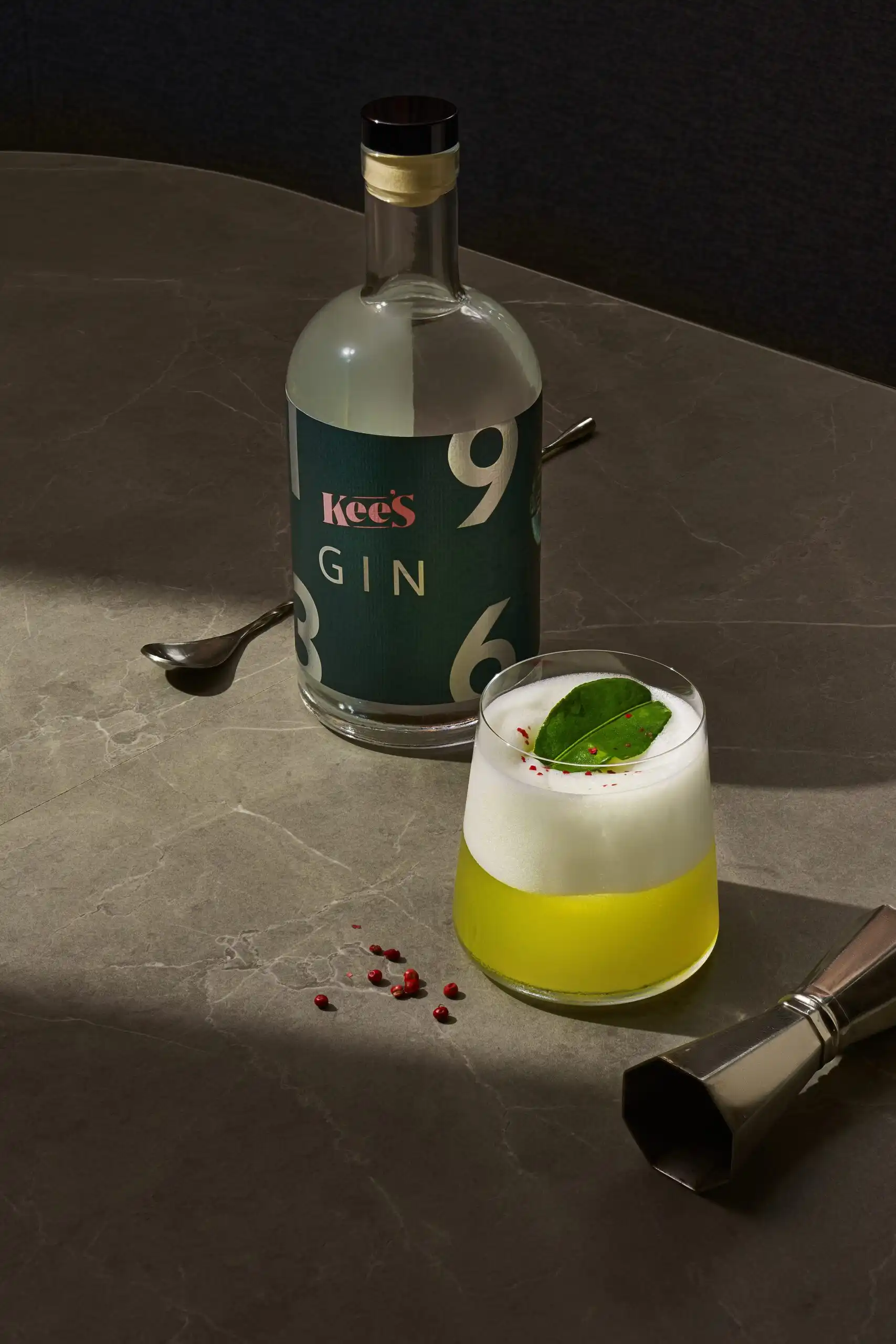 Kee’s gin bottle with a citrus cocktail topped with foam and leaf garnish on a marble table.