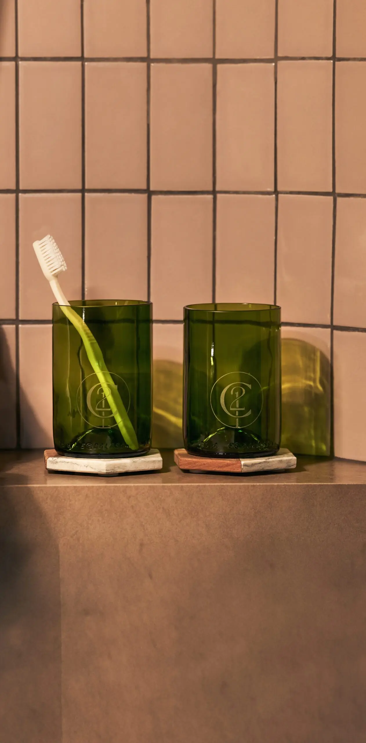 Green glass tumblers with toothbrush on a tiled bathroom counter with warm lighting.