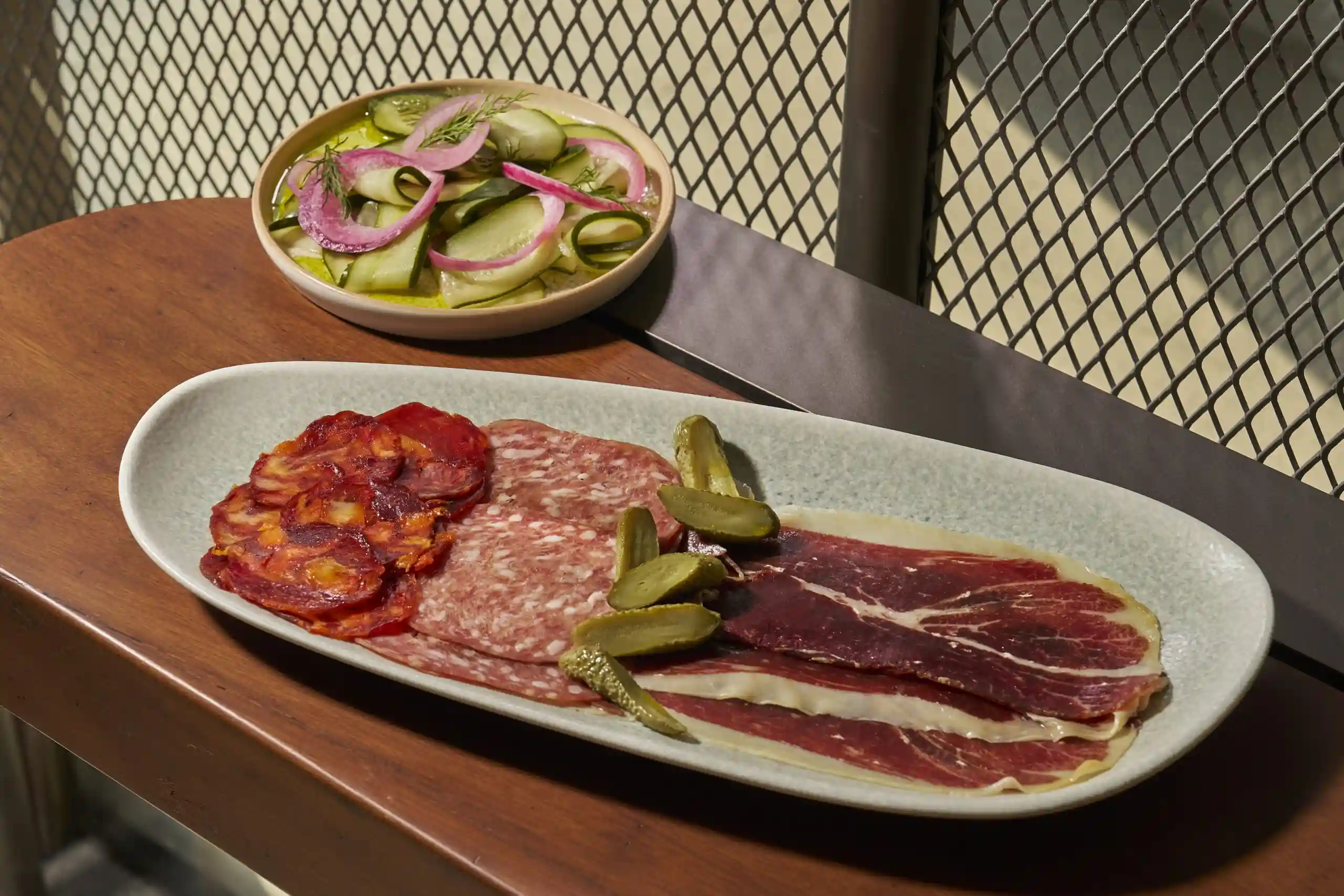 Charcuterie platter with cured meats, pickles and cucumber salad served on a wooden table.