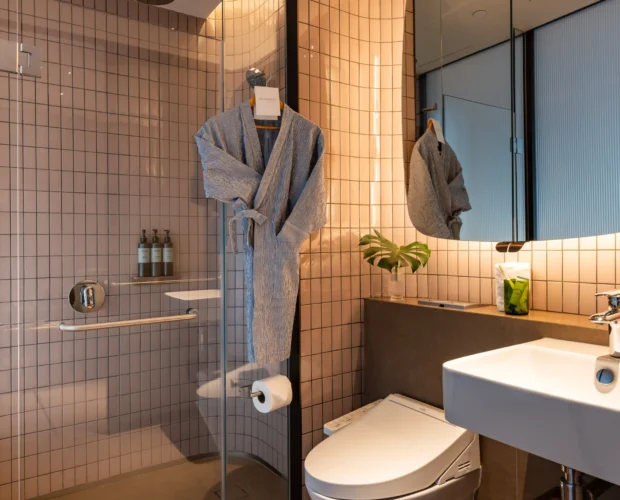 Bathroom at 21 Carpenter hotel in Singapore with a bathrobe on a hanger, mirror and shower in soft warm lighting